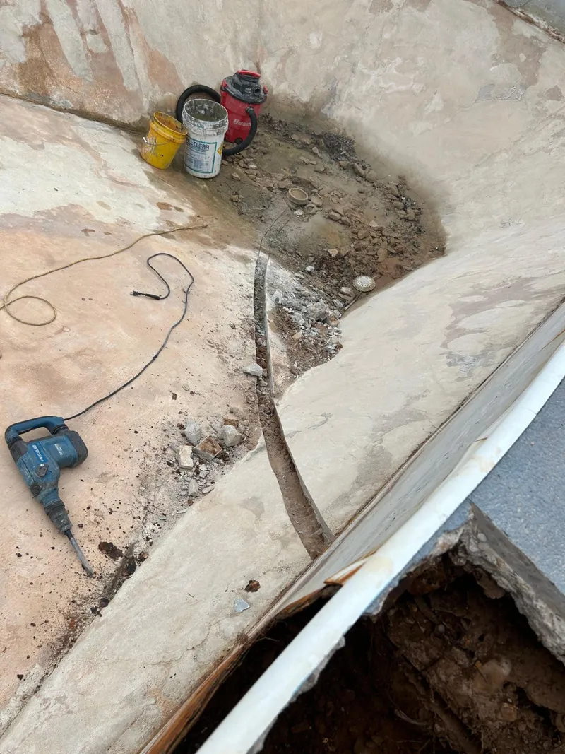 Pool floor cut and trenched from the deep end drain toward the wall, with a demolition hammer and buckets on the floor